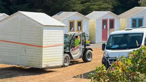The photo shows a row of timber beach huts on a dry, grassy area, with a small lifting vehicle carrying one of the huts while a white van is parked nearby. Several other huts remain standing in the background, painted white with brightly coloured doors and window frames, while trees form a dense backdrop behind the site.