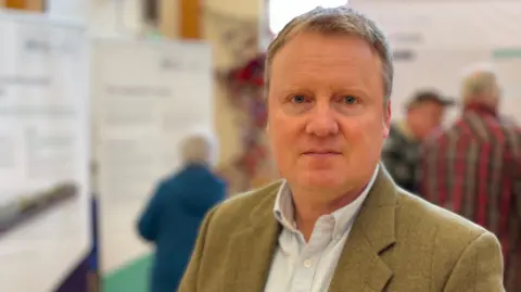 A man with short fair hair looks at the camera. He is wearing a light blue shirt and a brown-green jacket. He is standing in a room full of display boards and people looking at them. He is in focus while the background is out of focus.