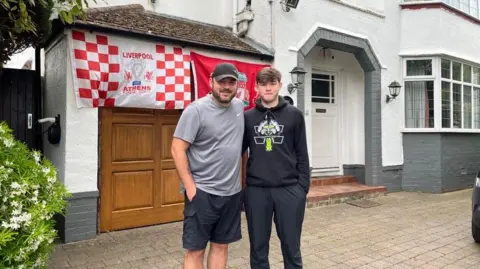 Jonny Humphries/BBC Phil Cross, 40, who has a grey t-shirt, baseball cap and dark blue shorts, stands next to his son who has a black hoodie and jogging bottoms. Behind them are two Liverpool flags hanging on the garage of their semi-detached house.