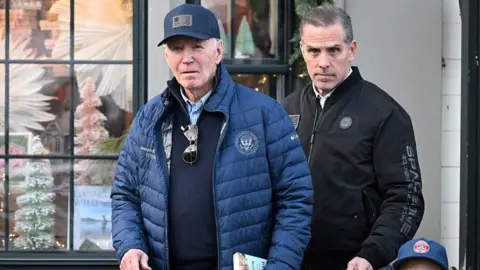 The president on the left wearing a blue coat and a baseball cap while Hunter Biden behind him is wearing a black jacket. They are in front of a window display.