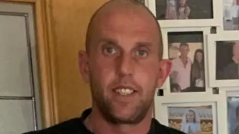 A man with short dark hair and a beard smiling at the camera wearing a black t-shirt.