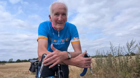 An older man with white hair is on a bicycle in a field of crops. He is wearing a blue cycling t-shirt that reads "hope house children's hospices"