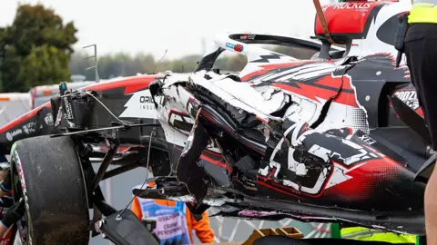 Getty Images A red, white and black F1 car being lifted up onto a trailer after a crash. The body work on the car is significantly damaged. Workers are helping to guide the car onto the trailer. 