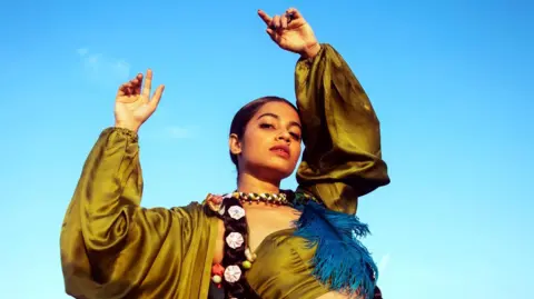 Tejasvi Patil Tejasvi Patil, strikes a pose with both arms in the air, wearing a green silk top decorated with a large blue feather 