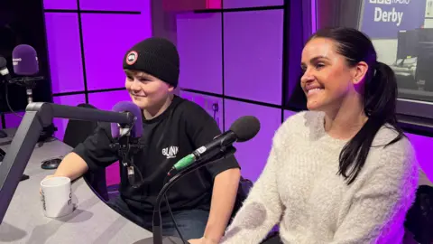 A boy and woman sit in a purple radio studio. The boy is on the left, wearing a black woolly hat and black t-shirt, both with white logos. The woman has long black hair tied back and a woolly white jumper. The pair are sat looking to the left in front of microphones. The boy is holding a mug of tea.