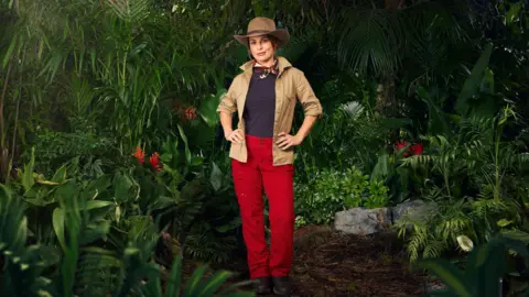 ITV Coleen Ronney wears a beigh flak jacket, red trousers and  a necktie and is pouting directly at the camera with her hands on her hips. In the background are jungle plants and red flowers. 