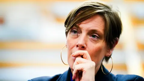 PA Jess Phillips wears large gold hoops, a blue blazer and holds her hand to her mouth. 