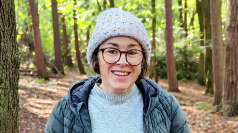 Jonny Coxon Charlotte stood in woodlands wearing glasses and a woolly hat