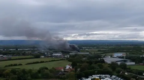 Green fields and hills in the background. In the centre of the image there are buildings and black smoke is rising from one of them. There are parked vehicles to the left of the aerial image. 