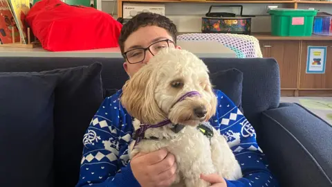 Cayden is sitting on a navy sofa. He is wearing a blue and white jumper, has black glasses on and short black hair. On his lap is a white dog with a harness around its outh. 