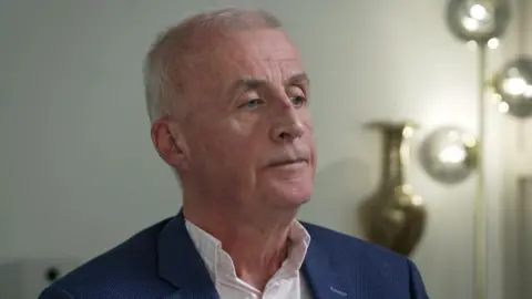 A man with short grey hair looks to the right of the camera. He is wearing an open neck shirt and navy blue blazer Behind him is a tall lamp, illuminating the dimly lit room. 