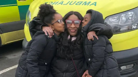 London Ambulance Service Carole crouches down in front of a yellow ambulance and hugs Pearl and Amelia who are both wearing padded black jackets.