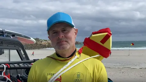 Close up of Alan Jenner at the beach. He is wearing a yellow rash vest and a blue cap. A red and yellow flag is on the beach behind him.