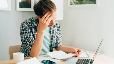 Man sits at a table with a laptop, a smartphone and a teacup in front of him, as he looks at bills.