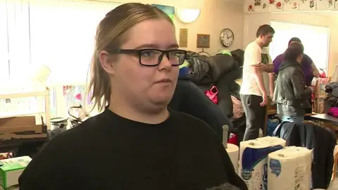 Jodie Holmes is has glasses and blonde hair pulled back into a ponytail. She is standing in the community centre. In the background, more people are sorting donations like kitchen roll and clothes.