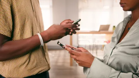 Two people stand facing each, looking down at their phones.