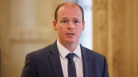 PA Media Communities Minister Gordon Lyons, with balding brown hair, wearing a white shirt, navy tie and navy suit. 