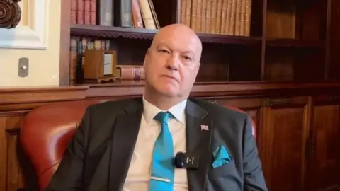 A bald man sitting in a dark red chair in front of dark wood panelling. He is wearing a suit with a white shirt and blue time. He has a union jack pinned to his lapel.
