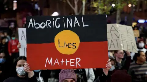 ABC/Jack Fisher A protestor holding up a sign that says Aboriginal Lives Matter painted on an Aboriginal flag, with another placard nearby with the partial words for Black Lives Matter