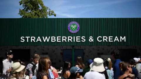 A crowd of people walk past a green building. The structure has a large sign that reads "strawberries and cream" and has the Wimbledon tennis logo.