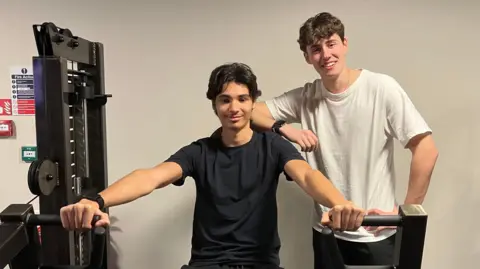 Marwan and Arthur in a gym, positioned at a strength-training machine. Marwan is seated and holding the machineās handles, while Arthur stands beside him with a hand resting on his shoulder. Both are wearing athletic clothing, and the gym equipment surrounds them in a well-lit workout space.