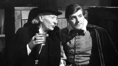 A black-and-white medium shot of two men sitting in a 16th century tavern set in a 1960s television studio. They are both in period dress. The man on the left is in his late 50s but made up to look older, with a long white wig and a black hat and black cloak. The man on the right is in his late 20s, also wearing a cloak, and a doublet with a ruffled collar.