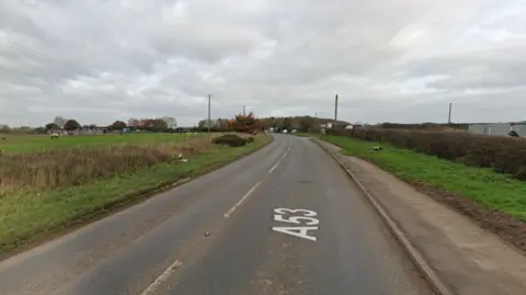 Google A Google Streetview screengrab of the crash site. A road stretches away into the distance, with A53 superimposed on to it. On the right hand side there's a pavement, and a grassy verge and hedgerow. On the left is a grassy verge with a short hedge with farmland visible beyond it. The sky is grey and overcast. There are bunches of flowers lying either side of the road - the tributes to both men who died. 