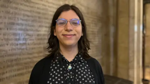 Head and shoulders of Sefira Davison. Sefira has shoulder length brunette hair, is wearing glasses and has a blue cardigan and black top with white shells on it.