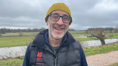 Mark pictured looking into the camera and smiling. He's wearing a navy blue knitted jumper, with a black gilet over the top. The jacket has a red, embroidered National Trust oak leaf logo on the right hand side, with 'National Trust Attingham Park' written on it. Mark is always wearing a yellow knitted hat and a pair of glasses. He's stood on a footpath, with the river visible just behind him. Grassland stretches off into the distance, with trees on the horizon - and the silhouette of the Wrekin just visible in the distance. The sky is grey and cloudy.