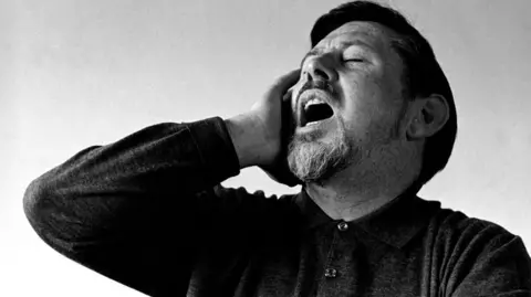 Getty Images A black and white photo of bearded singer Ewan MacColl, who holds his right ear as he sings loudly