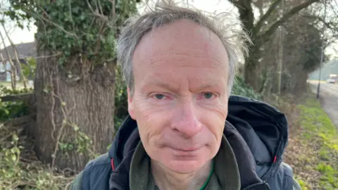 Tim Chapman is looking directly into the camera, while being photographed by the side of a road. He has grey, wispy hair, and is wearing a blue hooded jacket with a dark shirt underneath. In the background are trees.