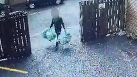 A man carrying four bags of rubbish, two in each hand. He is walking through an open gate into a driveway, with wooden fencing either side. His face is obscured. 