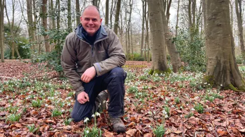 Charlotte Vowden/BBC A smiling Dan Winter, the head gardener at Evenley Wood Garden, kneels down on the forest floor amidst snowdrops that have pushed their way up through soil and dry leaves 