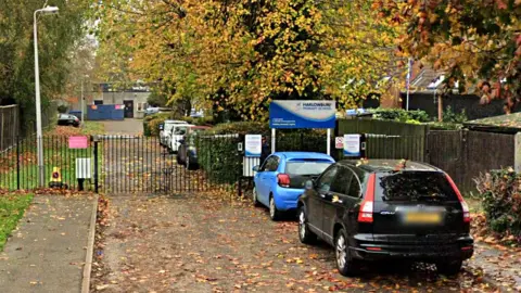 The entrance to Harlowbury Primary School, with closed black metal gates across the front of a driveway. The school building is distant in the background. A number of cars are parked to the right of the driveway and there are a number of leaves from trees on the ground. There is a sign for the school on the gates.