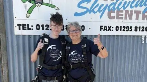 Sarah Allen Two people standing side by side in front of a metal wall with a large “Hinton Skydiving Centre” sign. Both are wearing matching dark charity T-shirts, parachute harnesses, and casual trousers with trainers. Each person is giving a thumbs-up gesture. The sign behind them includes a cartoon skydiver and visible contact details, indicating the location is a skydiving facility.
