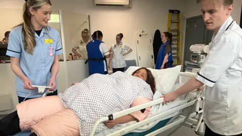 BBC A student led on a hospital bed as two medical students tend to her