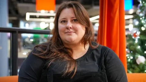 Picture of Esyllt Williams sitting on an orange sofa in an office. She is looking at the camera and has brown hair and eyes. She is wearing a black t-shirt.