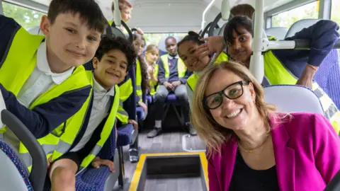 School children sat on a bus wearing their school uniform and a hi-vis jacket. The West of England Mayor is sat on the bus with the children. All are smiling and looking forward.