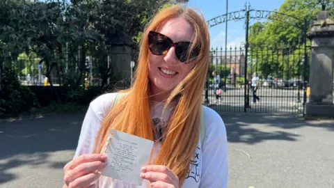 Mercedes has long, ginger hair. She is wearing tortoise-shell patterned sunglasses and is smiling at the camera. She is holding a small piece of paper in her hands with a typed up poem.