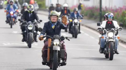A large group of motorcyclists ride down a road, with the front one waving at the camera.