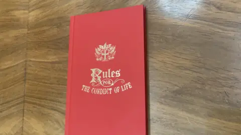 A red hardback notebook with gold lettering ready 'Rules for the Conduct of Life' lies on a polished oak table