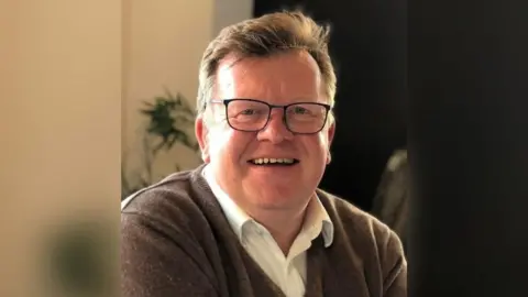 Chris Ratcliffe smiling and wearing glasses. He has a brown jumper on and a white collared shirt with the top button undone