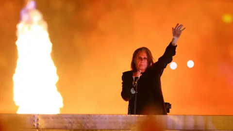 Osbourne with his left hand aloft performing on stage with microphone in his other hand. A pyrotechnic flares to his right, casting an orange glow behind him. He is wearing a black T-shirt and blazer, heavy eyeliner and his dark hair is long.