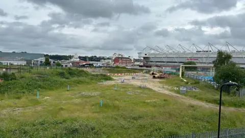 LDRS An empty plot of land with a few bits of debris littered across it and some sparse vegetation, with St Mary's stadium in the background