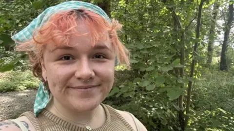 Holly Haines Selfie of Holly Haines, who has orange hair partially covered by a checked bandana. There are trees and foliage behind her.