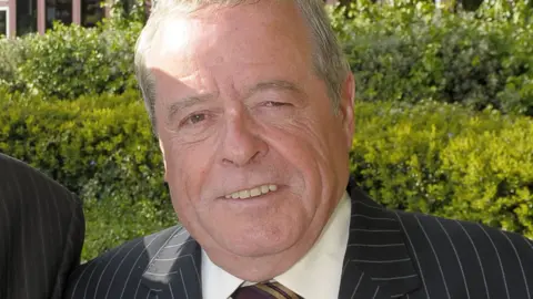 A man, who is prominent in the image and looking at the camera, is wearing a black and white striped jacket, a light shirt and a tie. Much of the background features bushes. 