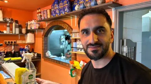Martin Heath/BBC Baris Danirt with short dark hair and beard smiling at the camera, standing behind the counter in a coffee shop. There are orange and blue walls behind him, and a serving hatch behind which another man is preparing an order. A spray bottle of disinfectant is in the foregound, and a further bottle of green cleaning fluid can be seen next to the hatch.