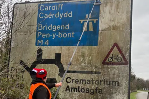 Clean Cymru Man in front of a road sign cleaning it with window cleaning equipment. He is stood next to a busy main road.