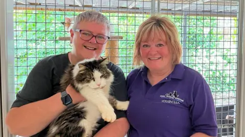 Stan is pictured in a rescue environment, being held by Elaine Brown and Sue Emmerson in front of a secure enclosure. The structured surroundings and visible signage indicate a formal animal care setting, while the close physical handling demonstrates attentive care and human interaction within the shelter. Elaine Brown has short white hair, glasses, dark top, loop earrings and a watch. Sue Emmerson has bobbed blonde hair and a purple top with a Eden Animal Rescue logo and the word "volunteer".