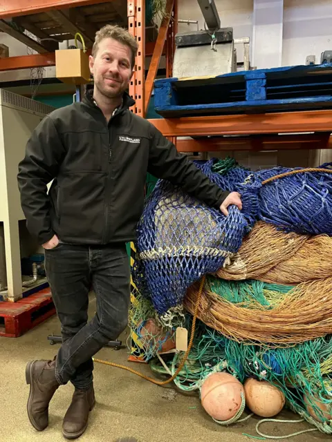 UHI A man with dark hair and a light beard in dark clothing stands beside fishing nets